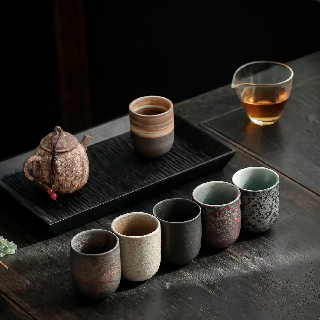 Set of ceramic tea cups and teapot on a dark wooden surface with a glass of tea.