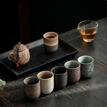 Set of ceramic tea cups and teapot on a dark wooden surface with a glass of tea.